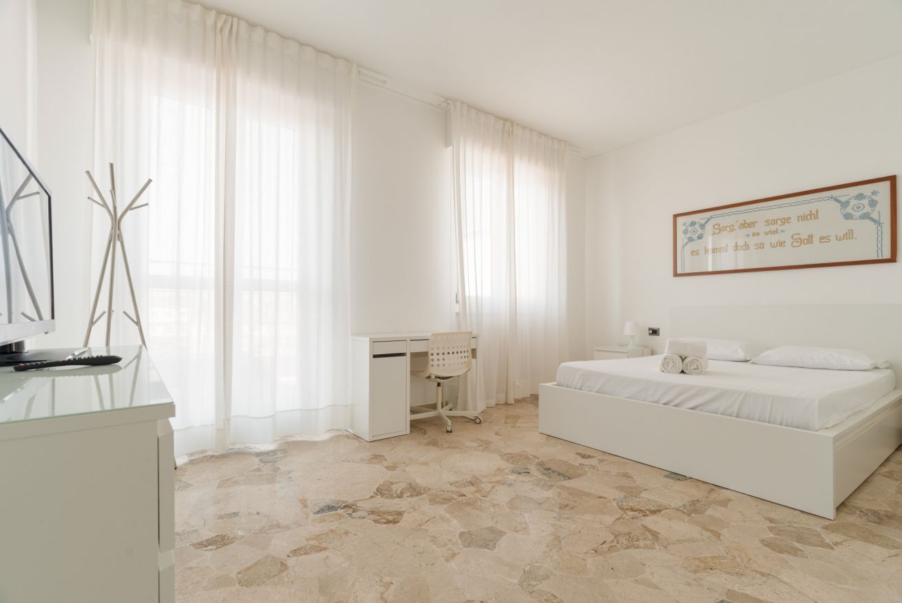 A bedroom with a bed, a desk, and a chair. There is a window with curtains, a wall-mounted artwork, and a stone-patterned floor.