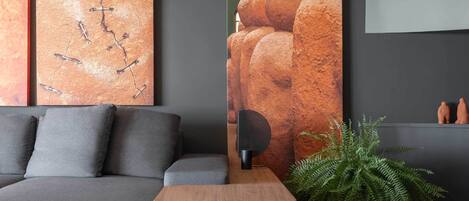 Una sala de estar moderna con un sofá gris, una mesita de madera, un cuadro abstracto grande, una planta de helecho y un altavoz.