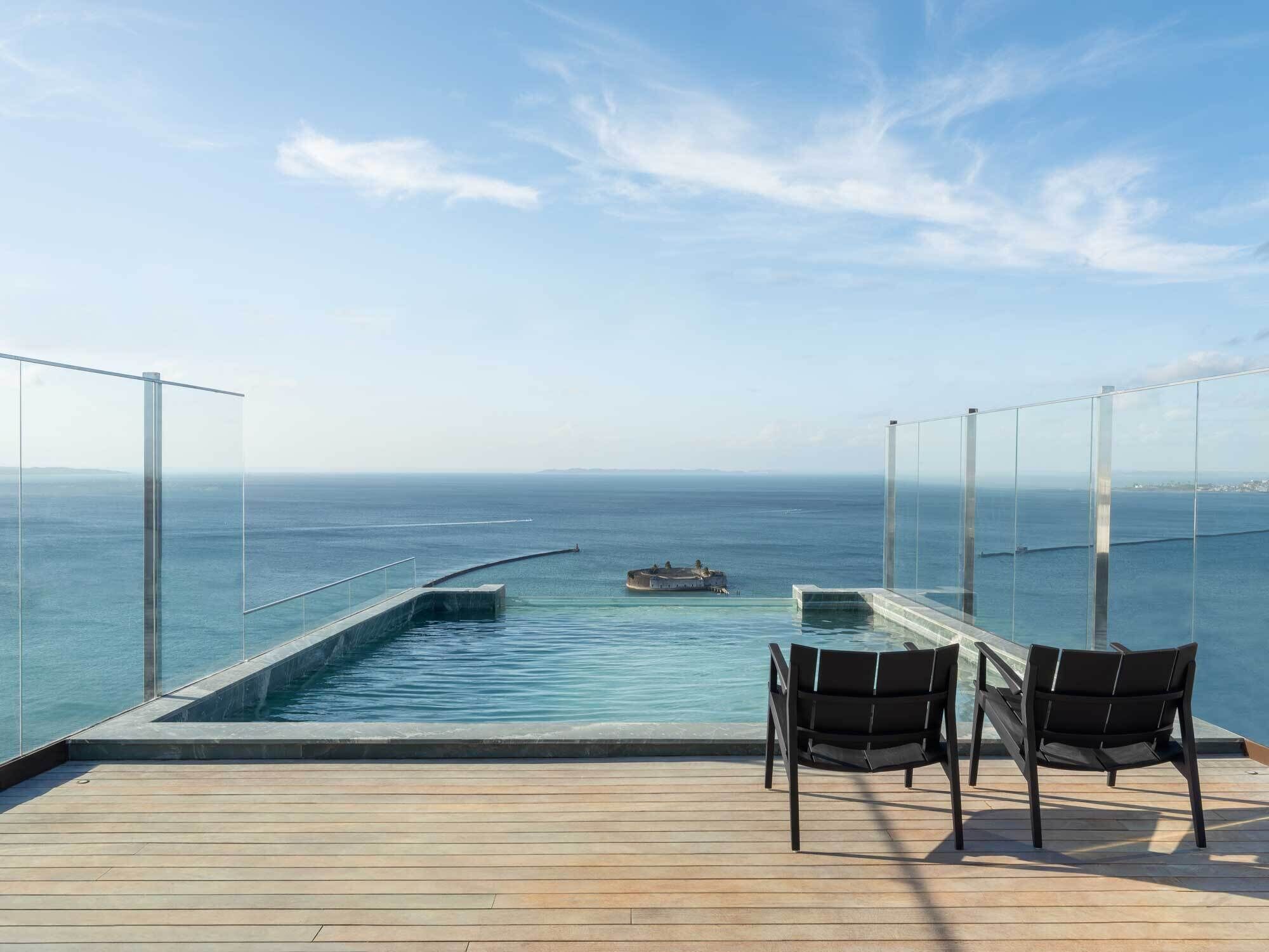 Uma área à beira da piscina com vista para o oceano, duas cadeiras e um barco ao longe.