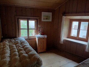 A cozy room with a bed, a small wooden cabinet, a desk with a lamp, and a window with a curtain.