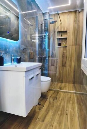 Ein modernes Badezimmer mit Holzboden, weißem Waschtisch, einer Duschkabine aus Glas und einem Holzregal.
