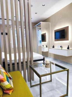 Ein modernes Hotelzimmer mit einem Glastisch, einer Couch mit farbigem Kissen, einem an der Wand befestigten Fernseher und einem großen Spiegel, der den Raum reflektiert.