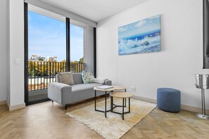 A modern living room with a grey sofa, a small round table, a blue ottoman, and a painting of a seascape. - One Rosebank 151 (Johannesburg)