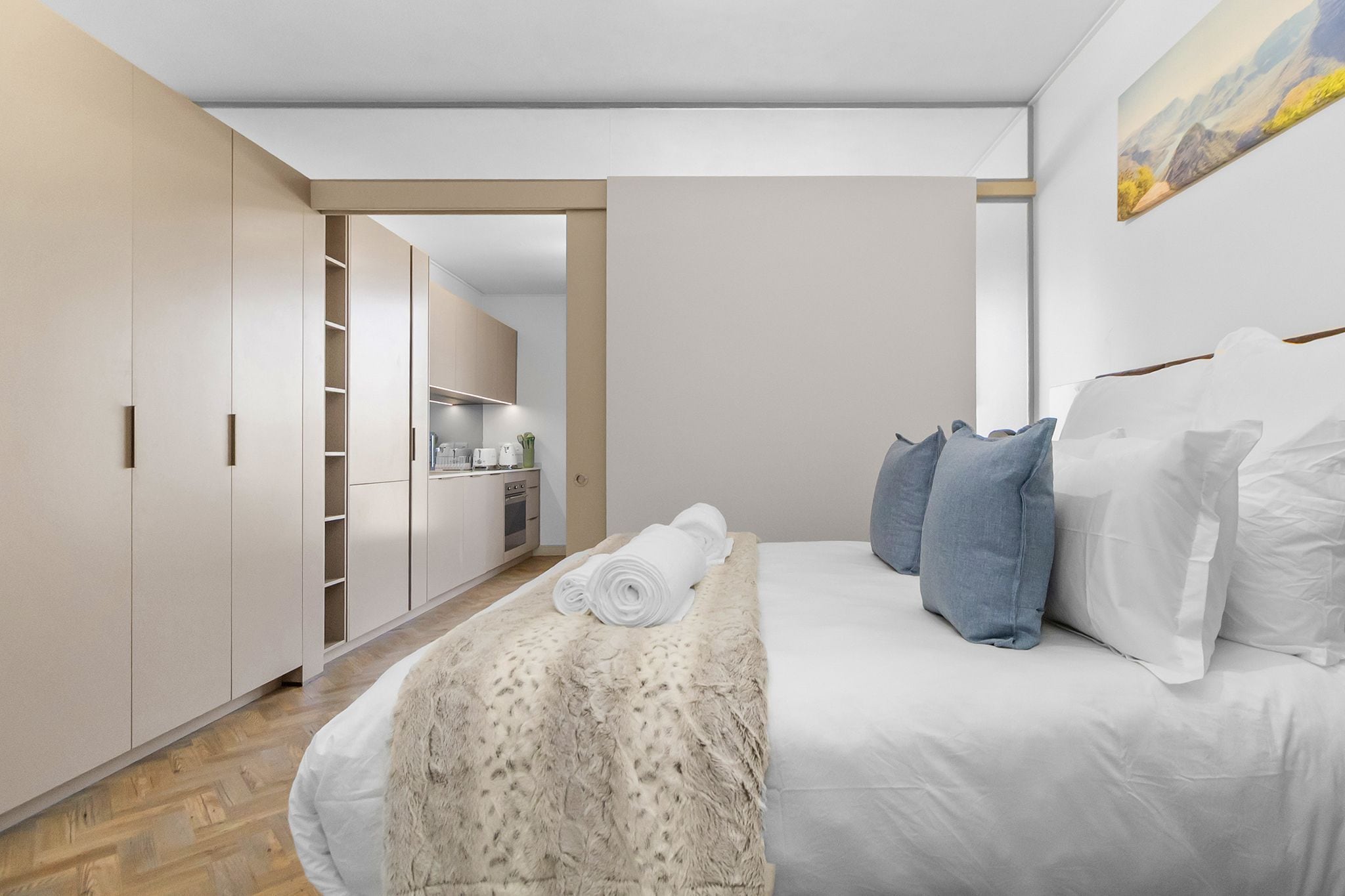 A bedroom with a bed, pillows, a rolled towel, a fluffy blanket, and a painting on the wall.