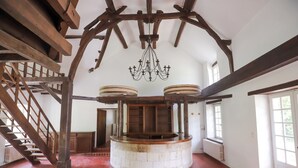 A room with a central circular stone structure, wooden beams, a chandelier, and a staircase.