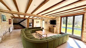 A spacious living area with a green sectional sofa, wooden beams on the ceiling, and a large window offering a view of the outdoors.