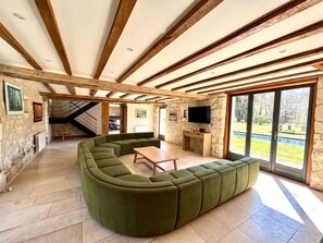 Ein geräumiger Wohnbereich mit einem grünen Ecksofa, Holzbalken an der Decke und einem großen Fenster mit Blick ins Freie.