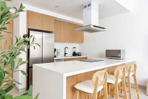 A modern kitchen with a central island, wooden cabinets, a stainless steel refrigerator, and a built-in range hood. - Coyote Tower 203 | Luxury Studio Walk to Marina (Cabo San Lucas)