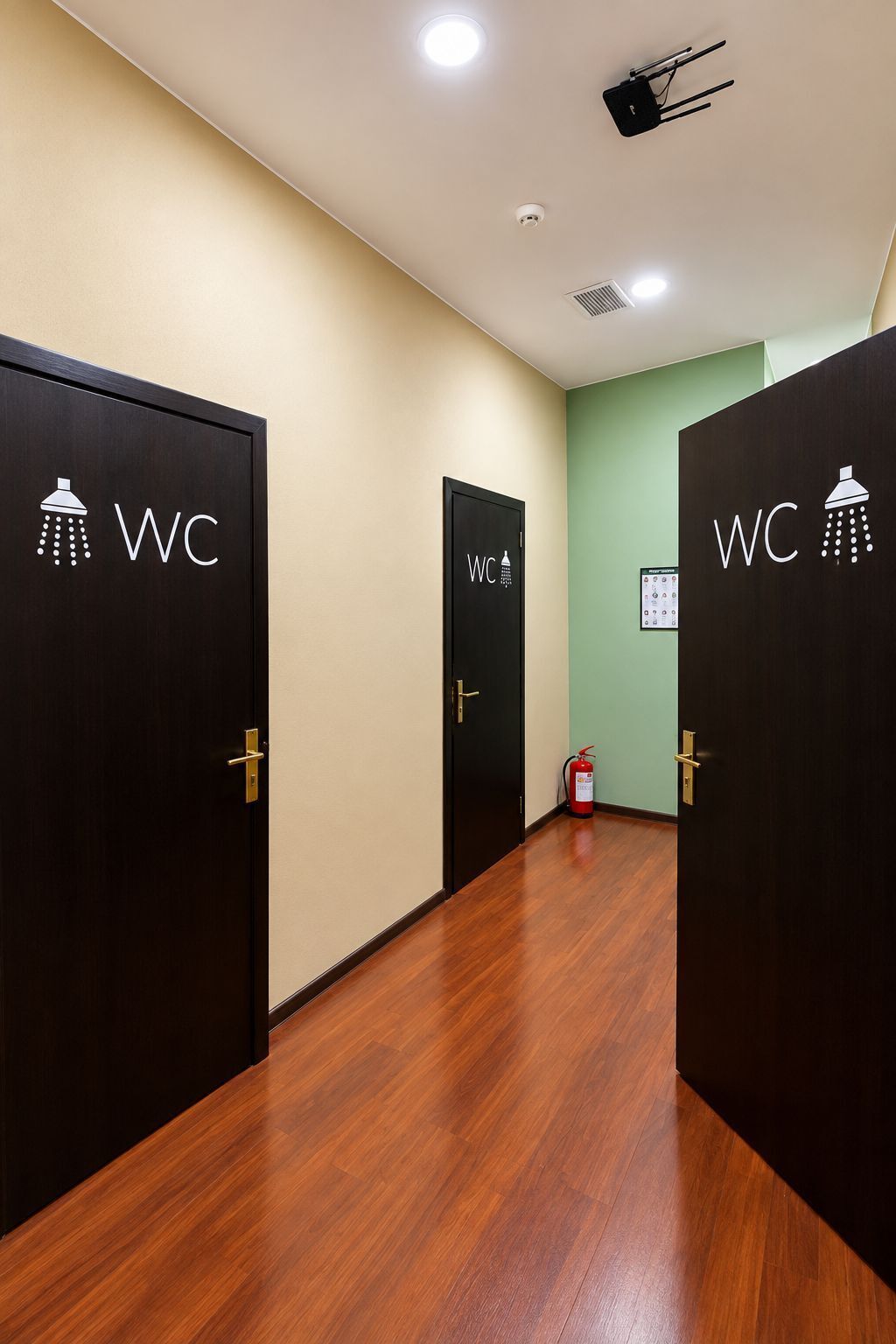Corredor com três portas identificadas como 'WC' para sanitários, um extintor de incêndio e um projetor fixado no teto.