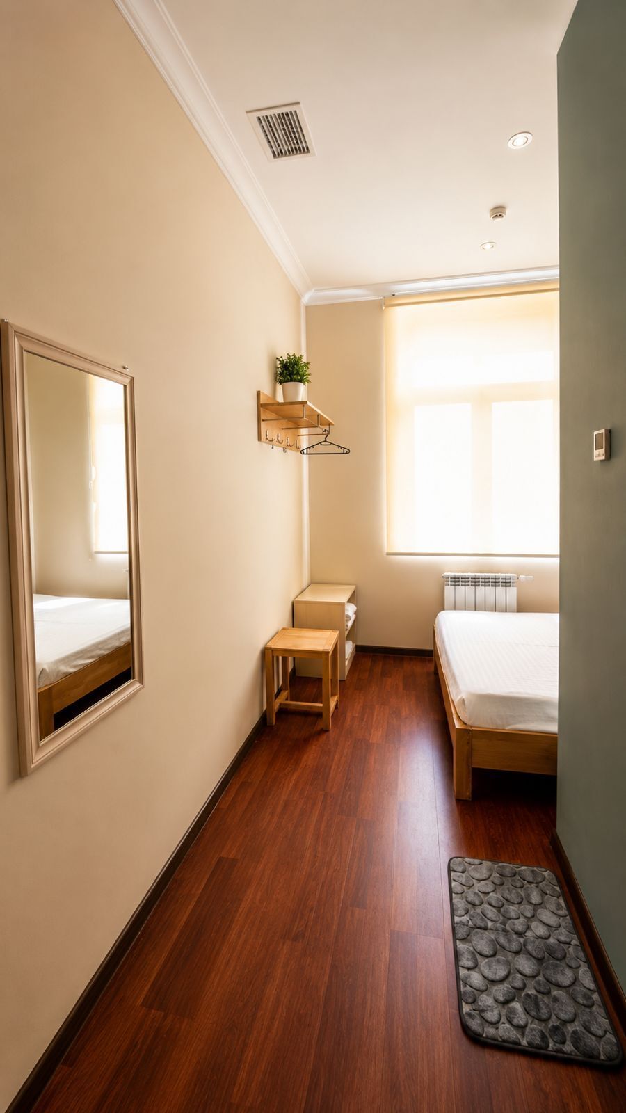 A hotel room with a wooden floor, a bed, a bedside table, a mirror, a radiator, and a window with blinds.