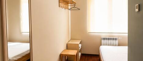 A hotel room with a wooden floor, a bed, a bedside table, a mirror, a radiator, and a window with blinds.