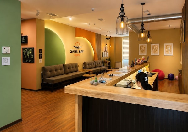 A modern hostel interior with a reception desk, seating area, and a wall-mounted sign for 'SAHIL BAY HOSTEL'.