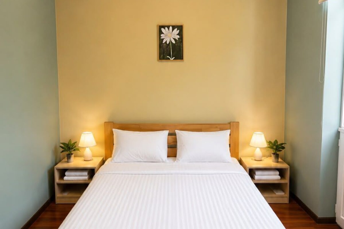 A neatly made bed with white linens in a room with wooden flooring and a single wall-mounted light fixture.