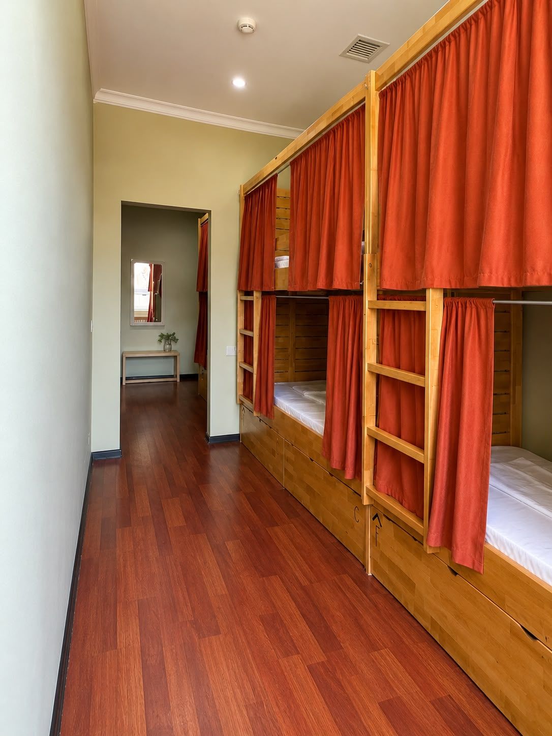 A room with bunk beds, wooden flooring, a mirror, and a small table.