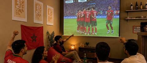 A group of people watching a sports event in a room with a large screen, a flag, and a shelf with bottles.