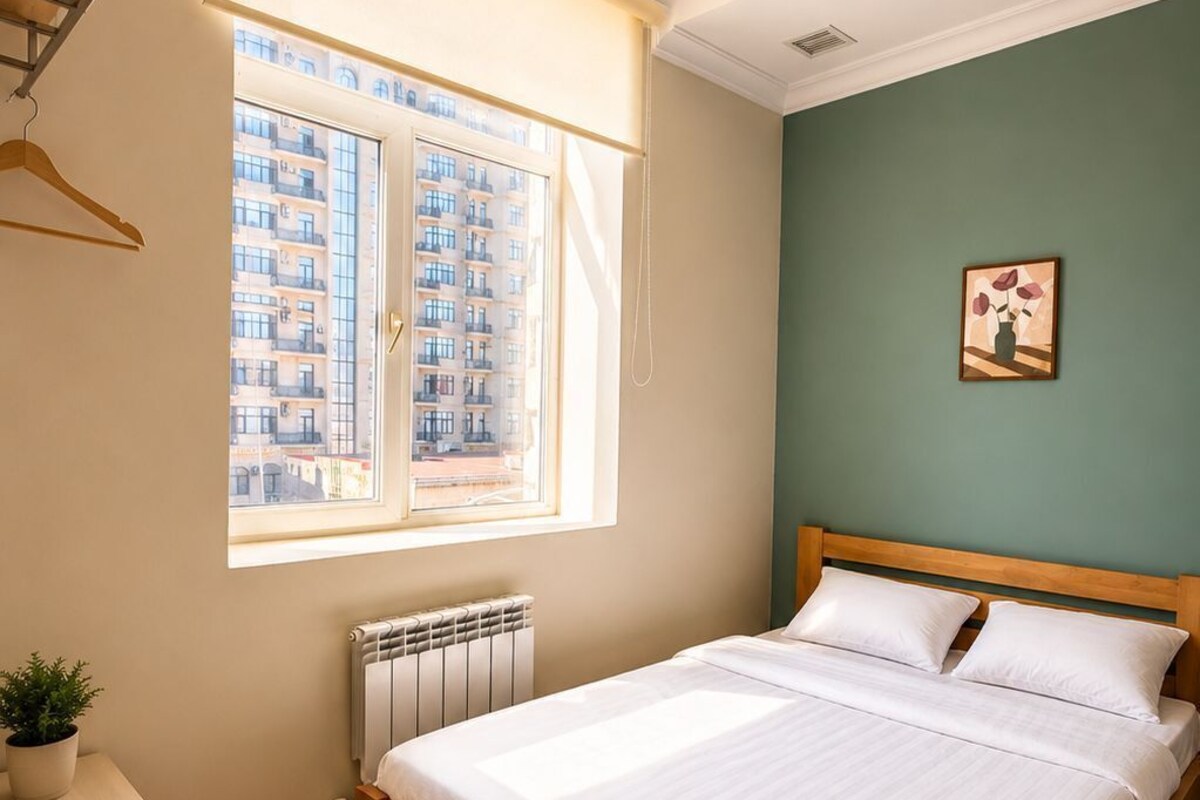 A bedroom with a bed, a radiator, a window with a view of buildings, a wooden table, and a wall-mounted picture.