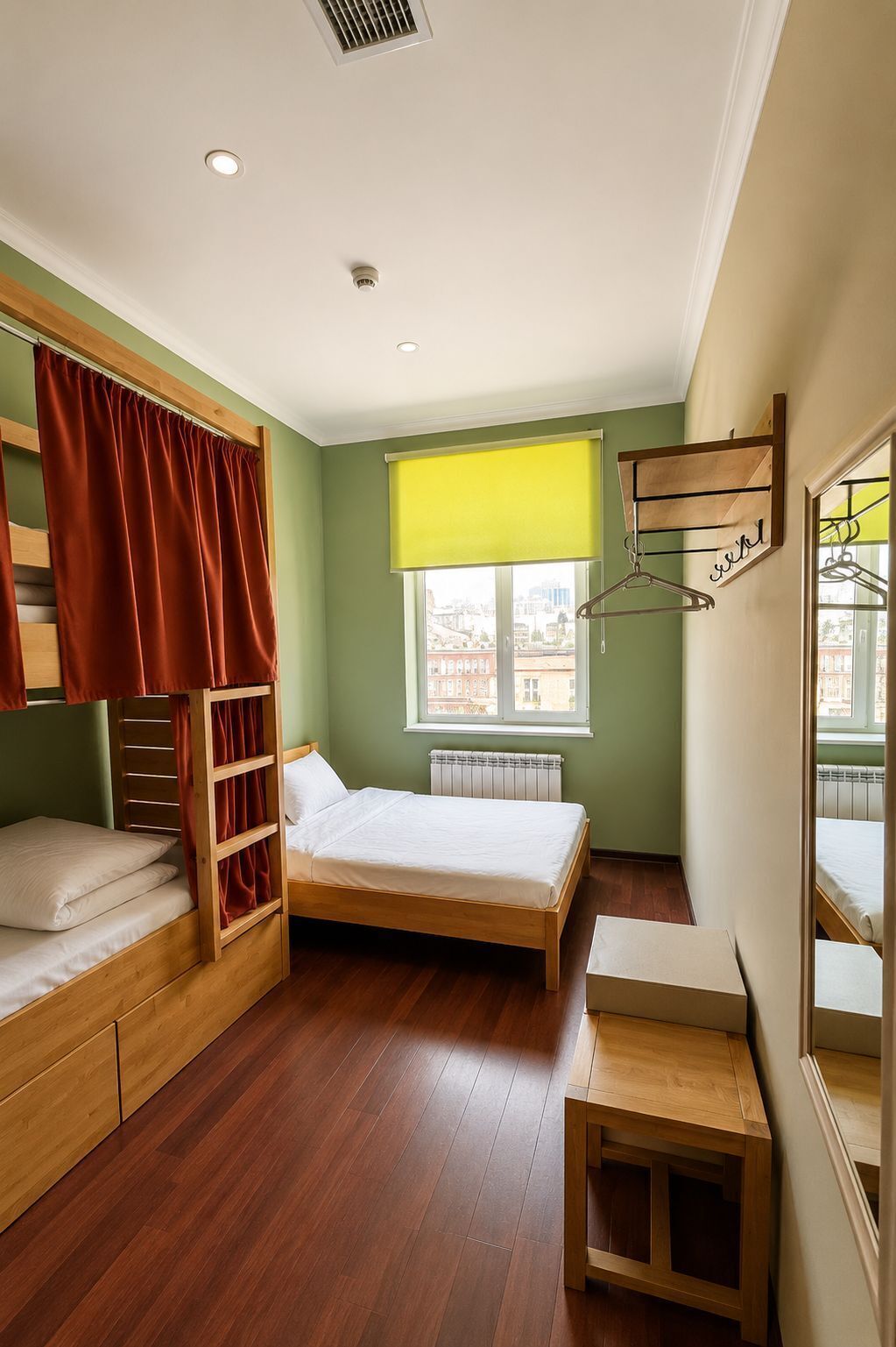 Una habitación con literas, una cama individual, una ventana con persiana amarilla, un radiador, una mesa de madera y un espejo en la pared.