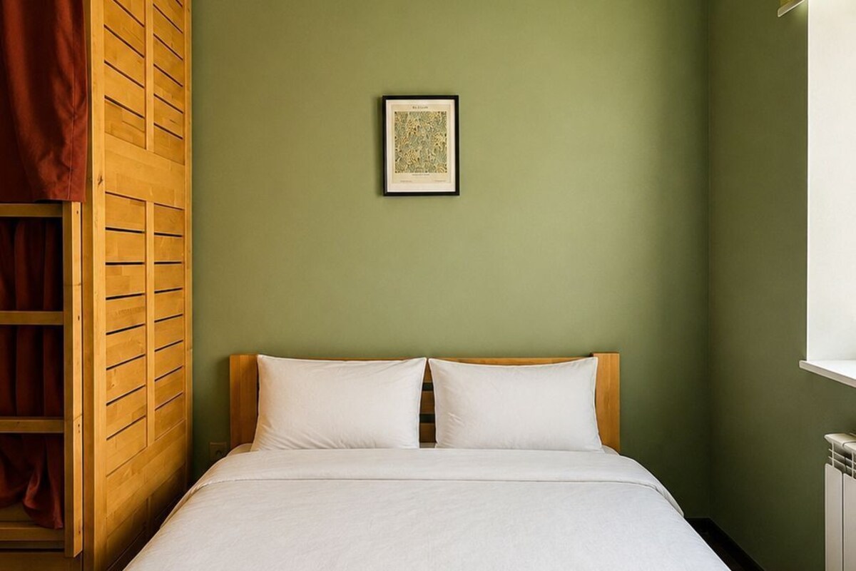A single bed with a wooden headboard, a framed picture on the wall, and a window with a yellow blind.