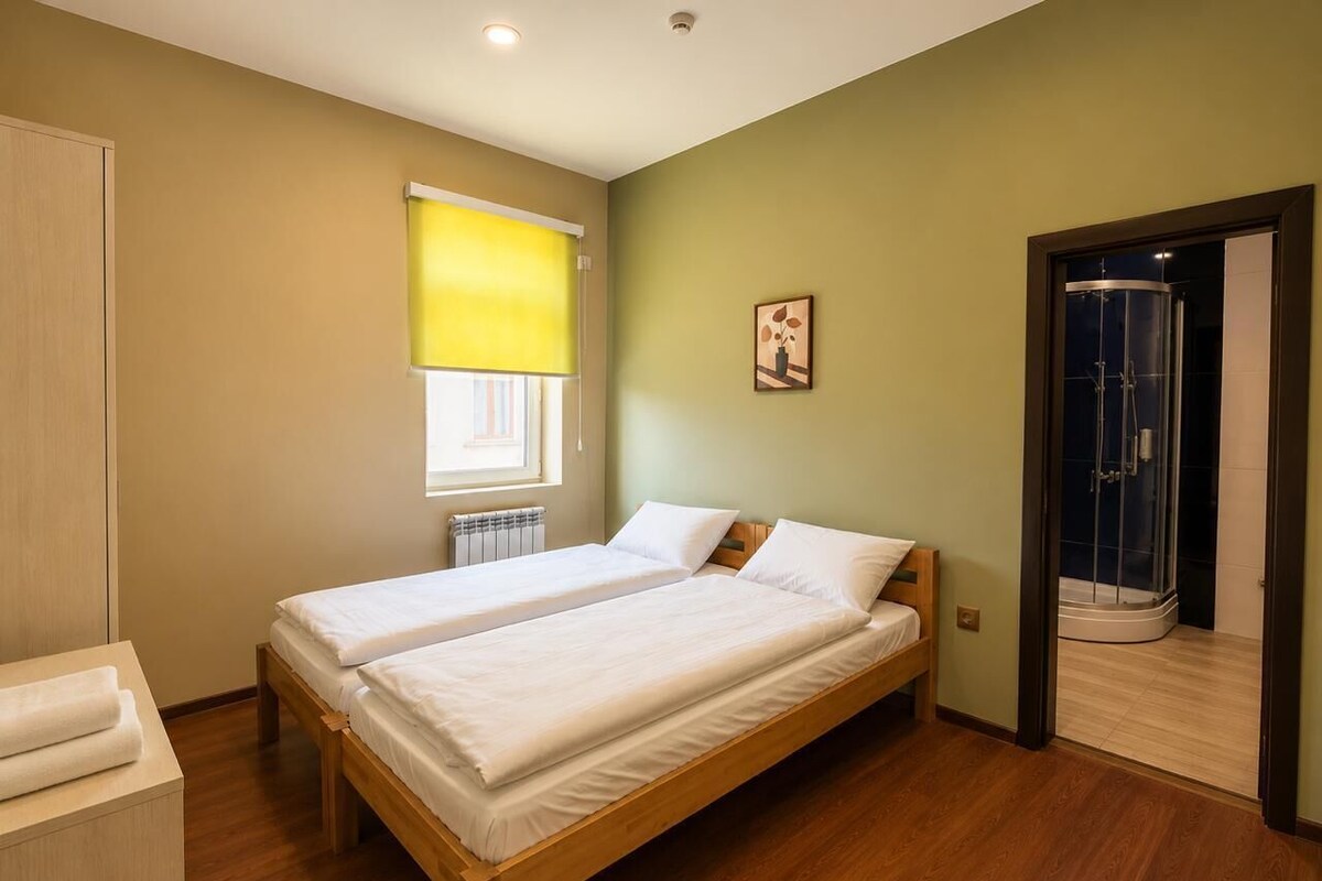 A room with two single beds, a wooden floor, a window with blinds, a radiator, and a shower area.