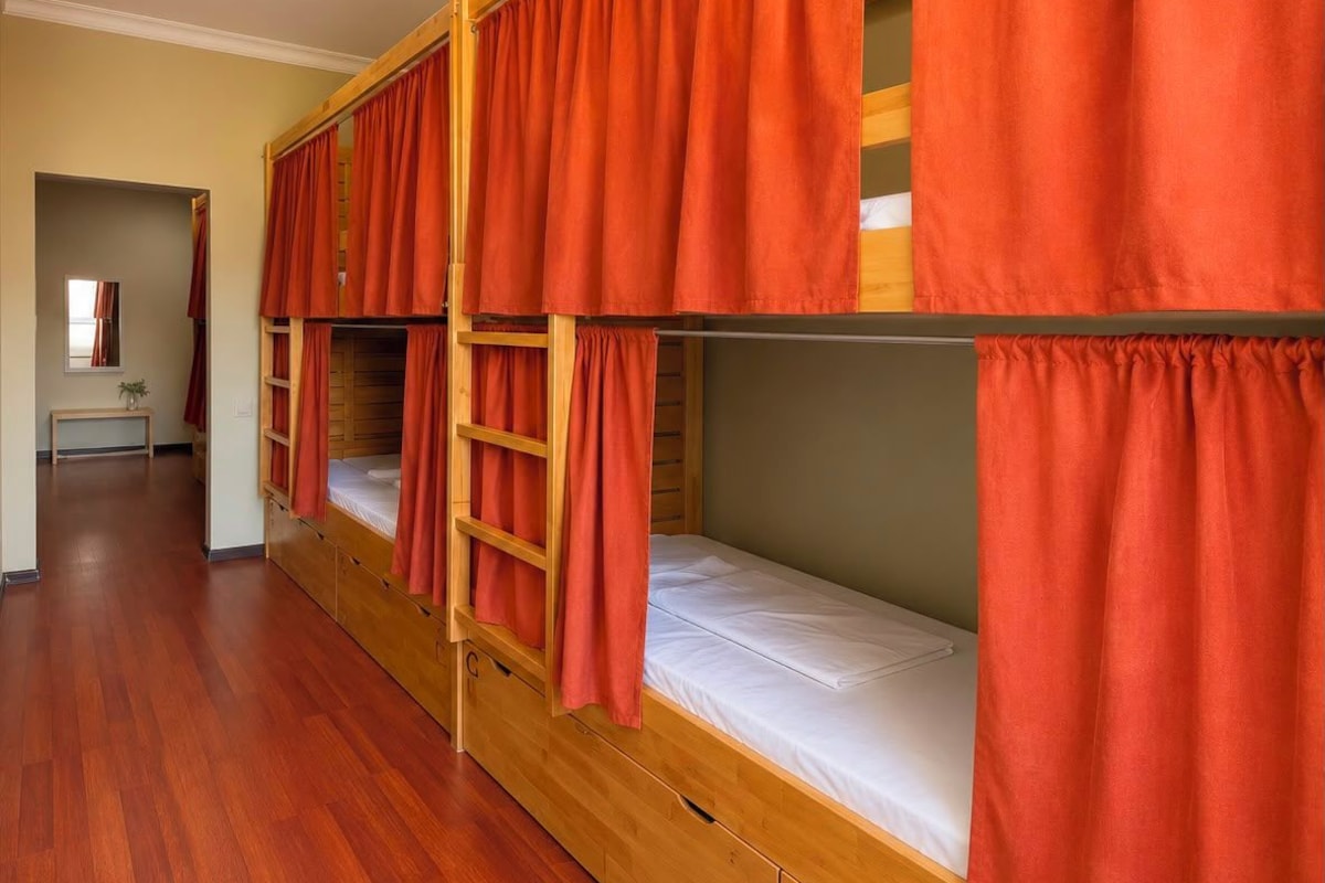 A dormitory room with bunk beds, wooden flooring, and red curtains.