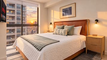 Une chambre d’hôtel avec un grand lit, une vue sur la ville, une œuvre d’art encadrée au mur et une table de chevet avec une lampe.