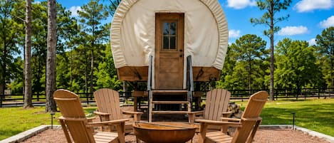 A covered wagon with a wooden door, surrounded by Adirondack chairs and a fire pit, set in a grassy area with trees.