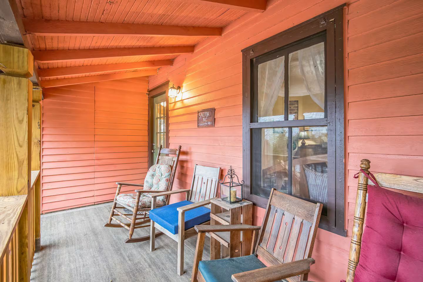 A wooden porch with two rocking chairs and a bench, a window with curtains, and a sign that reads 'SALUDA NE'.