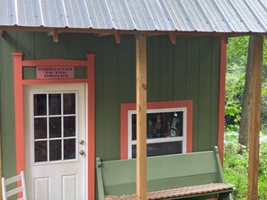 A small, rustic building with a green exterior and a red door. There is a bench outside and a window with a view of the surrounding greenery.