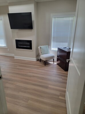 Una habitación con una televisión de pantalla plana montada en la pared, una chimenea debajo, un sillón blanco con cojín, un mueble de madera y una puerta que da a otra estancia.