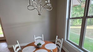 Een eetruimte met een ronde tafel, witte stoelen, een kroonluchter en een groot raam met uitzicht op groen.