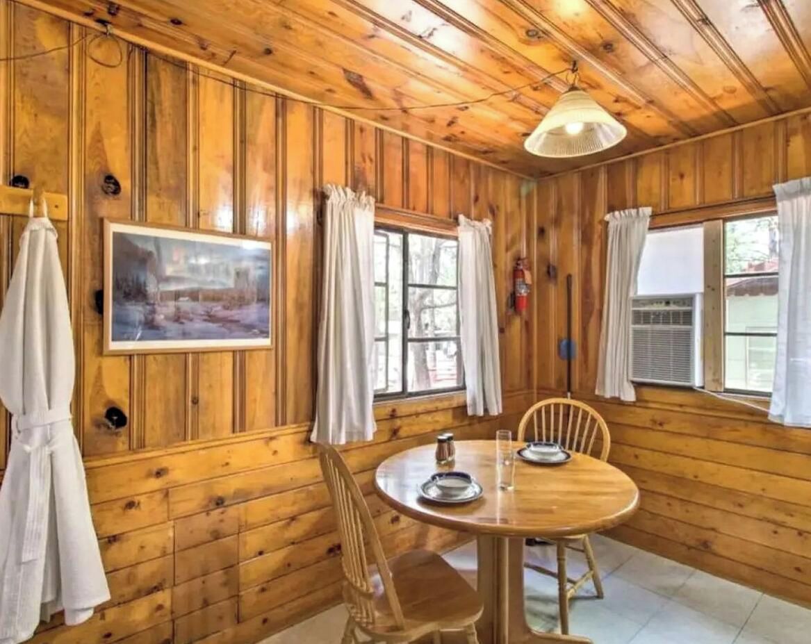 A wooden interior with a round table, chairs, a painting, and windows with curtains.