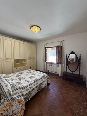 Ein Schlafzimmer mit Bett, Schrank, Fenster, Heizkörper, Spiegel und einem Korbwannenmöbel.