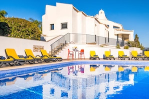 Ein Pool mit gelben Liegestühlen, ein weißes Gebäude mit Balkon und ein klarer, blauer Himmel.