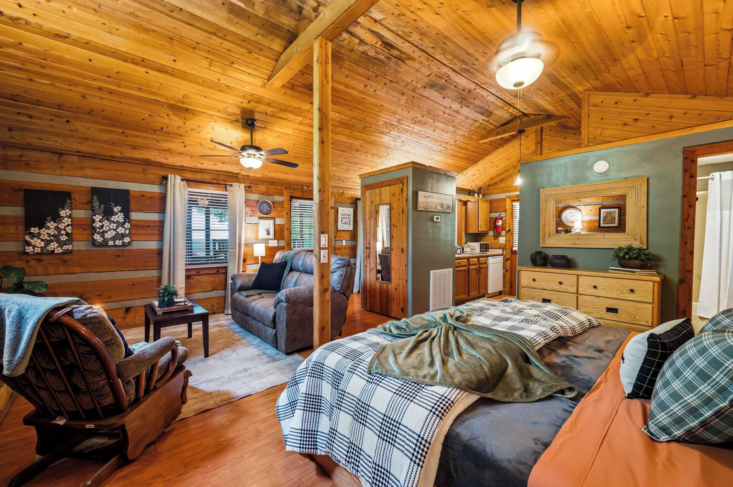 A cozy cabin interior with a wooden ceiling, a bed with a plaid blanket, a sofa, a rocking chair, and a dining area.