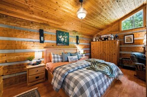 Ein gemütliches Schlafzimmer in einer Holzhütte mit Bett, Nachttischen, einer Nachttischlampe, einem Gemälde und einem Fenster mit Blick auf Bäume.