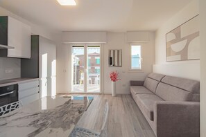 Ein modernes Wohnzimmer mit einer Couch, einem Essbereich mit Stühlen, einer Küche mit Herd und einer Glastür, die auf eine Terrasse im Freien führt.
