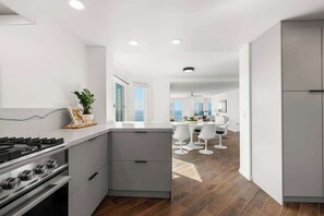 A modern kitchen with a central island, a dining area with white chairs, and a view of the ocean. - Oceanfront, AC, Fully Remodeled Unit (Solana Beach)