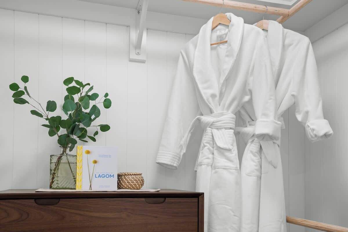 A white bathrobe hanging on a wooden hanger, a white vase with green leaves, and a small basket on a wooden dresser.