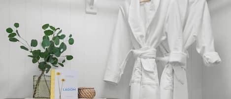 A white bathrobe hanging on a wooden hanger, a white vase with green leaves, and a small basket on a wooden dresser.
