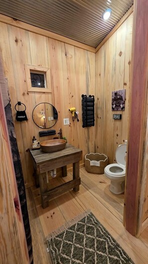 A wooden bathroom with a round mirror, a sink, a toilet, and a small window. - Cabin Retreat with Fire Pit & Scenic Ridge Views in Caddo Gap, Arkansas (Caddo Gap)