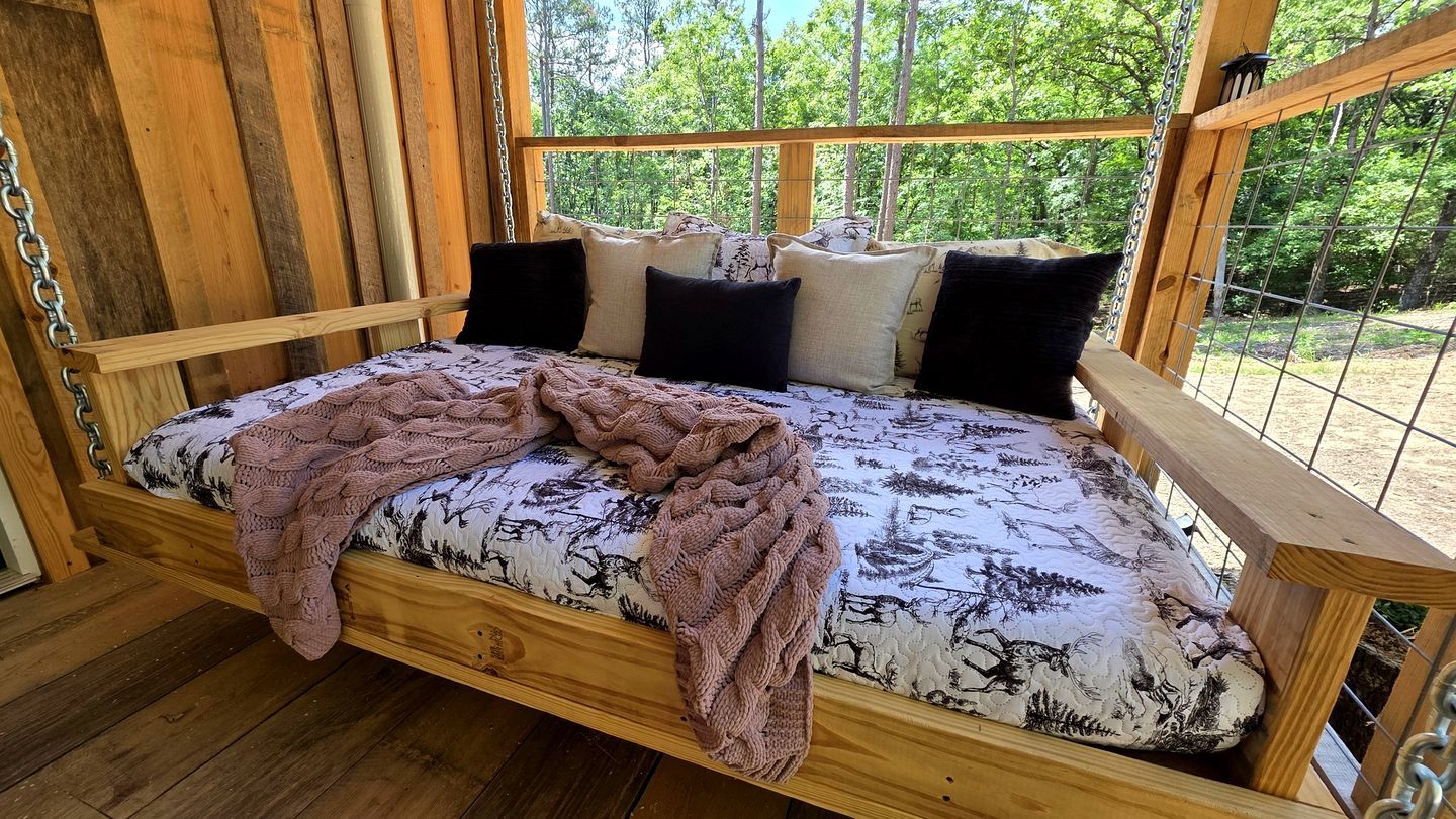A wooden bed with a patterned mattress, pillows, and a knitted blanket in a cabin with a view of trees.