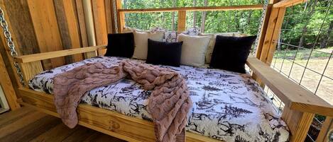 A wooden bed with a patterned mattress, pillows, and a knitted blanket in a cabin with a view of trees.