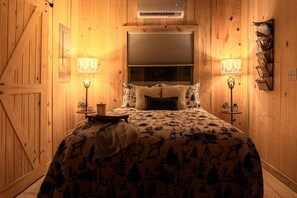 A cozy bedroom with a bed, two bedside lamps, a heater, and a wooden door. - Cabin Retreat with Fire Pit & Scenic Ridge Views in Caddo Gap, Arkansas (Caddo Gap)