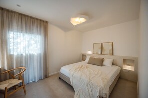 Ein Schlafzimmer mit einem Bett, einem Sessel, einem Fenster mit Vorhängen und Wandleuchten.