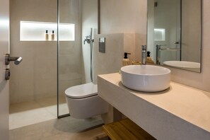 Een moderne badkamer met een douchecabine van glas, een wit toilet en een rond wastafeltje met een spiegel erboven.