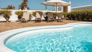 A swimming pool with lounge chairs and an umbrella, surrounded by neatly trimmed hedges and a white wall. - Villa Playa Vista Lobos (Corralejo)