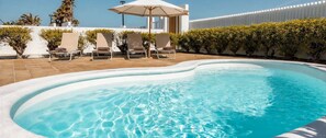 Ein Swimmingpool mit Liegestühlen und einem Sonnenschirm, umgeben von ordentlich geschnittenen Hecken und einer weißen Mauer.