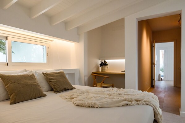 Un dormitorio con cama, almohadas, una mesita de noche con una planta y una silla.