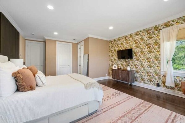 A bedroom with a large bed, a dresser, a flat-screen TV on the wall, and a window with curtains.