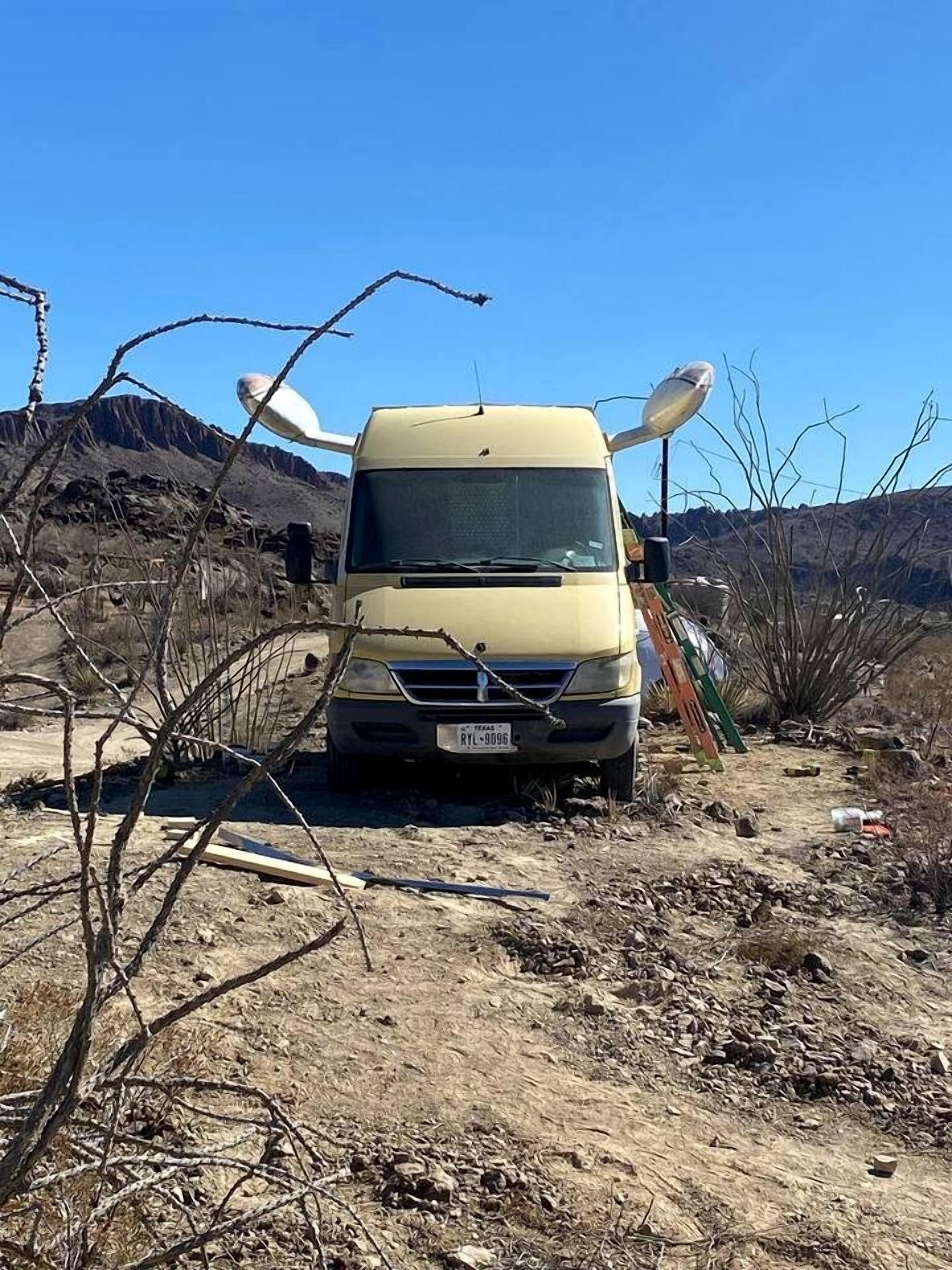 Un véhicule jaune est garé sur un chemin de terre, dans un paysage où la végétation est clairsemée et où l’on aperçoit des montagnes en arrière-plan.